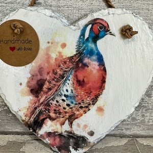 May include: Handmade heart-shaped slate plaque with a watercolor painting of a pheasant. The plaque has a brown twine hanger and a brown paper tag that says "Handmade with love".