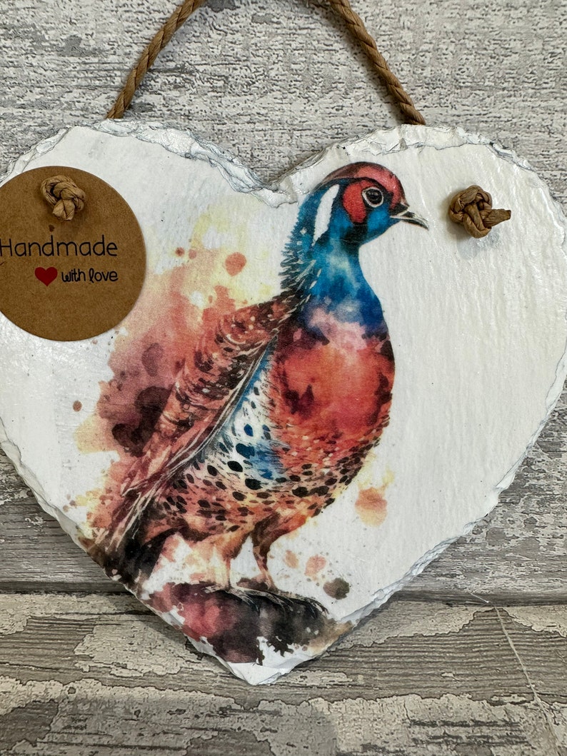 May include: Handmade heart-shaped slate plaque with a watercolor painting of a pheasant. The plaque has a brown twine hanger and a brown paper tag that says "Handmade with love".