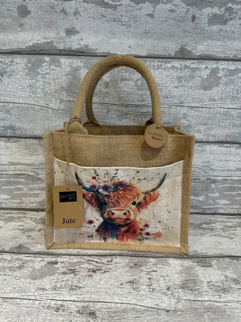 K&ouml;nnte beinhalten: Eine kleine braune Jute-Tragetasche mit einem Aquarellbild eines Highland-Rinds mit einer Blumenkrone. Die Tasche hat ein braunes Etikett mit der Aufschrift "Handmade with love". Die Tasche hat auch ein braunes Etikett mit der Aufschrift "Wisteria Mill Jute".