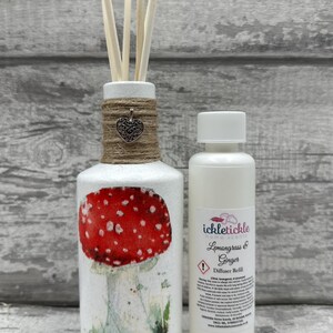 May include: White ceramic diffuser bottle with a red and white mushroom design and a jute twine bow with a silver heart charm. The bottle is filled with diffuser sticks. A clear glass bottle with a white label that reads "ickletickle home scents Lemongrass & Ginger Diffuser Refill" is next to the diffuser bottle.