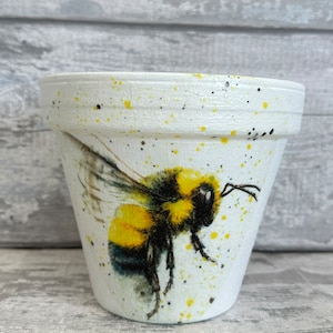Könnte beinhalten: Weißer Keramik-Blumentopf mit einer Illustration einer gelben und schwarzen Biene. Der Topf hat eine gesprenkelte weiße Glasur mit gelben und schwarzen Farbstippen.