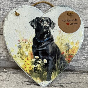 May include: A heart-shaped slate plaque with a black Labrador Retriever sitting in a field of flowers. The plaque has a brown circle with the text "Handmade with Love" on it.