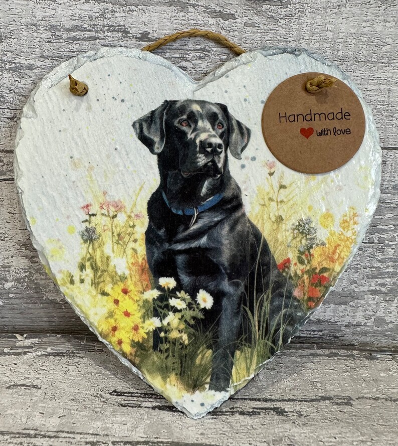 May include: A heart-shaped slate plaque with a black Labrador Retriever sitting in a field of flowers. The plaque has a brown circle with the text "Handmade with Love" on it.