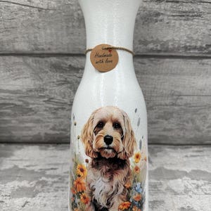 May include: White ceramic vase with a brown tag that says "Handmade with love". The vase features a floral design with a brown and white dog in the centre.