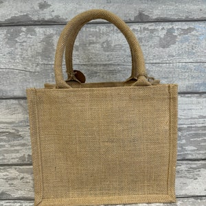 K&ouml;nnte beinhalten: Eine kleine, naturfarbene Jute-Tragetasche mit einem einzigen Henkel. Die Tasche ist aus gewebtem Stoff und hat ein schlichtes, rustikales Design.