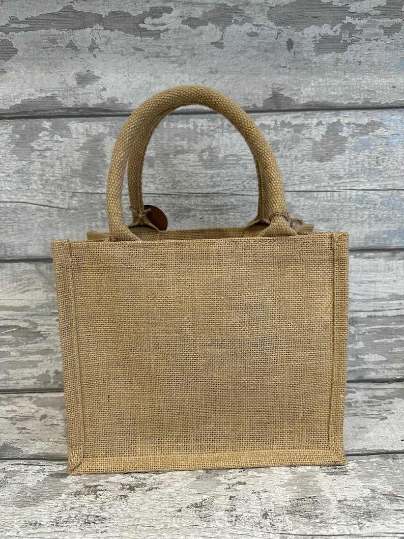 K&ouml;nnte beinhalten: Eine kleine, naturfarbene Jute-Tragetasche mit einem einzigen Henkel. Die Tasche ist aus gewebtem Stoff und hat ein schlichtes, rustikales Design.