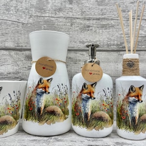 May include: A white ceramic bathroom set featuring a fox design. The set includes a tumbler, a vase, a soap dispenser, a diffuser bottle with reeds, and a small bottle. Each piece has a fox illustration and the words "Handmade with love".