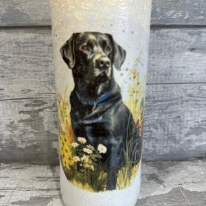 May include: White glass bottle with a black Labrador Retriever image. The dog is sitting in a field of yellow and white flowers. The bottle has a textured surface.