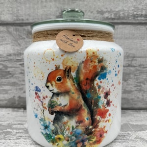 Könnte beinhalten: Weiße Keramikdose mit Glasdeckel, verziert mit einem Aquarellbild eines Eichhörnchens, umgeben von bunten Blumen. Die Dose hat ein braunes Etikett mit der Aufschrift "Handgefertigt mit Liebe".