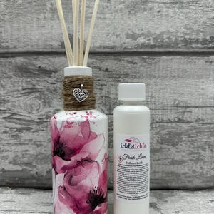 Peut inclure: Un diffuseur décoratif avec un motif floral rose et blanc, accompagné d'une bouteille de recharge. Le diffuseur a des bâtonnets en bois et une ficelle avec un charme en forme de cœur. La bouteille de recharge indique "Fresh Linen".