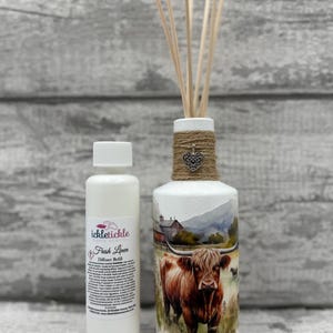 Peut inclure: Un flacon diffuseur blanc avec un motif de vache Highland, des détails en ficelle et des roseaux en bois. Une petite bouteille blanche avec le texte "Fresh Linen" est à côté. Le diffuseur est conçu pour parfumer une pièce.