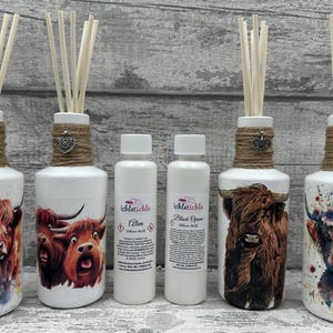 Peut inclure: Quatre bouteilles de diffuseur en céramique blanche avec de la ficelle brune et des breloques en forme de cœur argentées. Chaque bouteille présente une peinture à l'aquarelle d'une vache des Highlands. Deux bouteilles ont des vaches brunes et blanches, une a une vache noire et une a une vache brune avec une couronne florale. Deux petites bouteilles blanches avec des étiquettes qui indiquent "Tickle Tickle" et "Alien" et "Black Opium" sont au centre.