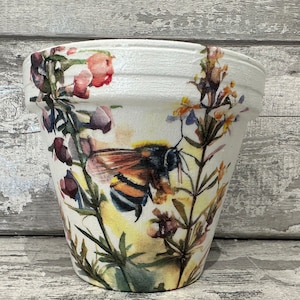 May include: White ceramic flower pot featuring a watercolour design of a bee and wildflowers. The bee is yellow and black with brown wings. The flowers are pink, purple and yellow.