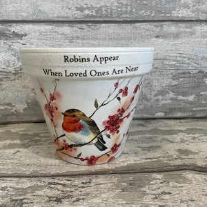 May include: White ceramic flower pot with a robin bird and red berries painted on it. The pot has the text "Robins Appear When Loved Ones Are Near" written on it.