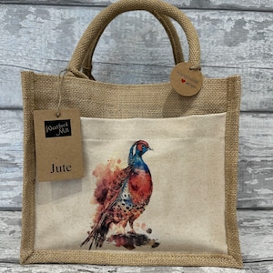 Könnte beinhalten: Eine braune Jute-Tragetasche mit einer weißen Stofftasche, die ein Aquarellbild eines Fasanen zeigt. Die Tasche hat ein braunes Etikett mit der Aufschrift "Westford Mill Jute" und ein rundes braunes Etikett mit der Aufschrift "Handgefertigt mit Liebe".