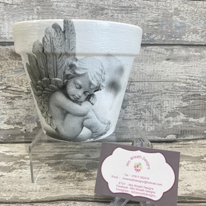 Könnte beinhalten: Ein weißer Keramik-Blumentopf mit einem grau-weißen Bild eines schlafenden Engels mit Flügeln. Der Topf steht auf einem klaren Acryl-Ständer. Eine Visitenkarte von Mrs. Wreath Designs ist ebenfalls auf dem Bild zu sehen. Die Karte enthält die Kontaktdaten des Unternehmens, einschließlich Telefonnummer, E-Mail-Adresse und Social-Media-Handles.