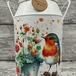 Könnte beinhalten: Eine weiße Milchkanne aus Metall mit einem Rotkehlchen und Blumen darauf bemalt. Die Kanne hat zwei Henkel und ein braunes Etikett mit der Aufschrift "Handmade with love".