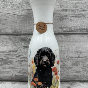 May include: White ceramic vase featuring an illustration of a black dog and wildflowers. The vase has a brown tag that reads "Handmade with love".