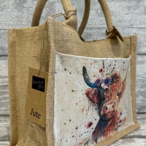 K&ouml;nnte beinhalten: Eine braune Jute-Tragetasche mit einer Fronttasche, die ein Aquarellbild einer Highland-Kuh mit einer Blumenkrone zeigt. Die Tasche hat zwei braune Jutegriffe und ein braunes Papieretikett mit der Aufschrift "Westford Mill Jute".