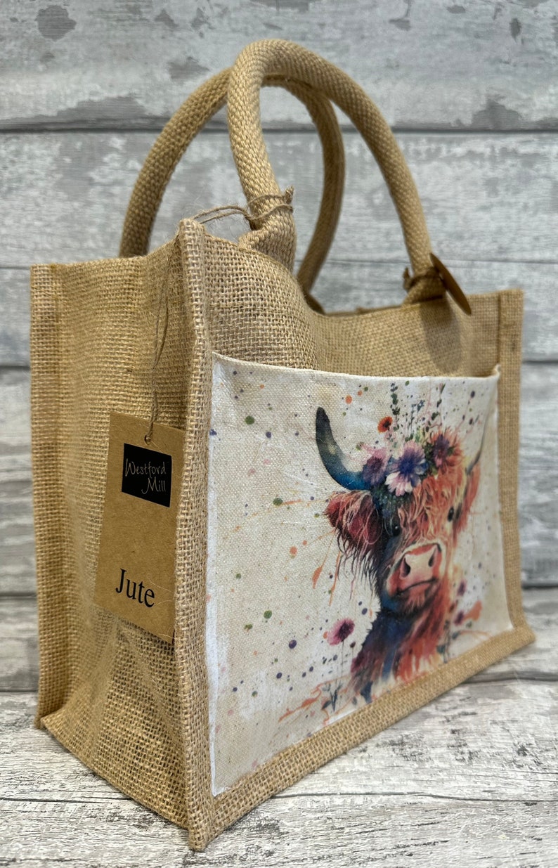 K&ouml;nnte beinhalten: Eine braune Jute-Tragetasche mit einer Fronttasche, die ein Aquarellbild einer Highland-Kuh mit einer Blumenkrone zeigt. Die Tasche hat zwei braune Jutegriffe und ein braunes Papieretikett mit der Aufschrift "Westford Mill Jute".
