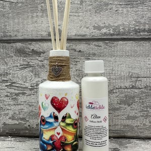 Könnte beinhalten: Eine weiße Diffuserflasche mit einer braunen Kordelschleife und einem silbernen Herzanhänger. Die Flasche hat eine bunte Illustration von zwei Fröschen, die ein rotes Herz halten. Die Flasche steht neben einer weißen Flasche mit einem Etikett, auf dem "ickletickle Home Scents Alien Diffuser Refill" steht.