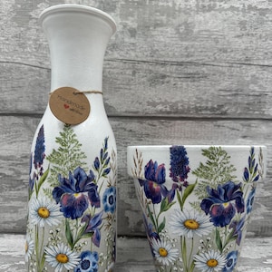 Könnte beinhalten: Weiße Keramikvase und passender Topf, verziert mit einem Blumendesign mit blauen Iris, weißen Gänseblümchen und grünem Laub. Die Vase hat ein Etikett mit der Aufschrift "Handmade with love".