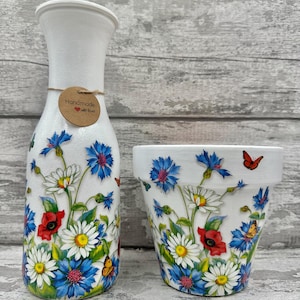 May include: A white ceramic vase and a matching flower pot are decorated with a floral design. The design features blue cornflowers, white daisies, red poppies, and orange butterflies. The vase has a narrow neck and a round tag that says "Handmade with love."