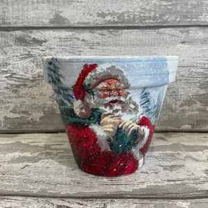 Könnte beinhalten: Ein weißer Keramiktopf mit einer festlichen Weihnachtsszene, die den Weihnachtsmann in einem roten Anzug vor einem verschneiten Hintergrund zeigt.