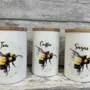 Könnte beinhalten: Drei weiße Keramikdosen mit Bambusdeckeln und Bienenillustrationen. Die Dosen sind mit "Tee", "Kaffee" und "Zucker" beschriftet.