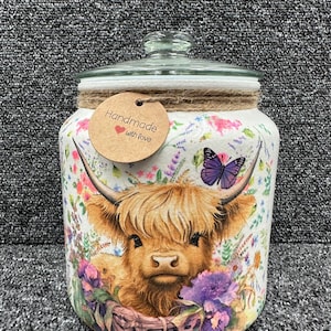 May include: A white ceramic jar with a glass lid decorated with a Highland cow, flowers, and a butterfly. The jar has brown twine tied around the lid and a brown tag that says "Handmade with love".