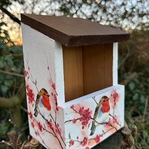 May include: A white birdhouse with a brown roof, decorated with a floral design and two robins. The birdhouse is painted with a watercolour-style design of pink flowers and red berries.