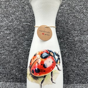 May include: White ceramic vase featuring a ladybug design and a brown tag that reads "Handmade with love".