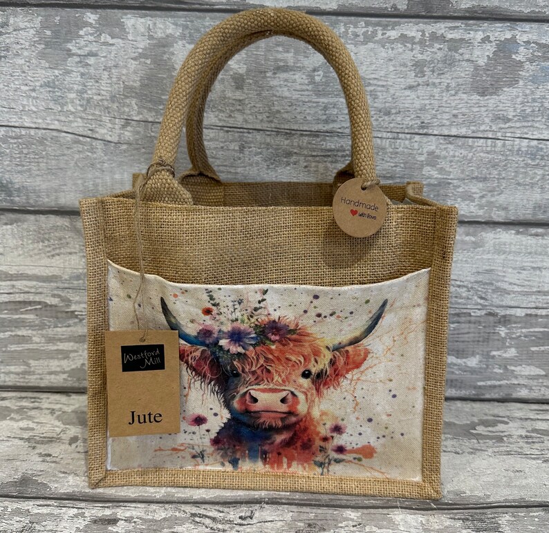 K&ouml;nnte beinhalten: Eine braune Jute-Tragetasche mit einem Blumenmuster einer Highland-Kuh. Die Tasche hat eine Tasche auf der Vorderseite und ein braunes Etikett mit der Aufschrift "Handmade with love".