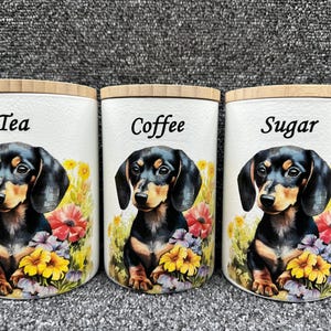 May include: Three white ceramic canisters with wooden lids and dachshund dog illustrations. The canisters are labelled "Tea", "Coffee", and "Sugar".