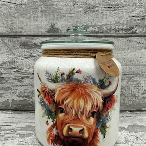 May include: White ceramic jar with a glass lid and a brown twine bow. The jar features a colourful illustration of a Highland cow with a floral wreath around its head. The jar has a brown paper tag with the text "Handmade with love".