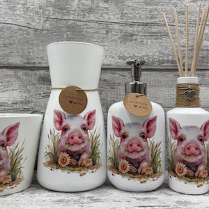 Könnte beinhalten: Eine Sammlung weißer Keramik-Badaccessoires mit Schweinedesign. Enthält einen Becher, eine Vase, einen Seifenspender, einen Diffusor und eine kleine Flasche. Jedes Teil hat eine Aquarellillustration eines Schweins.