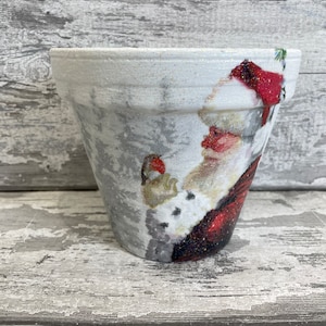May include: A white ceramic planter with a glittery finish features a vintage illustration of Santa Claus in a snowy scene. The planter is ideal for adding a festive touch to your home decor.