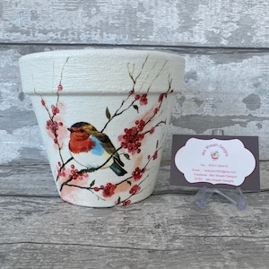 May include: White ceramic flower pot with a hand-painted design of a robin perched on a branch with pink blossoms. The pot is 6 inches in diameter.