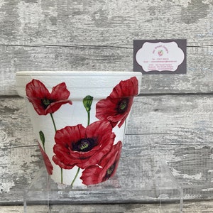 Könnte beinhalten: Weißer Keramik-Blumentopf mit rotem Mohnblumen-Blumendesign. Der Topf steht auf einem klaren Acryl-Ständer.