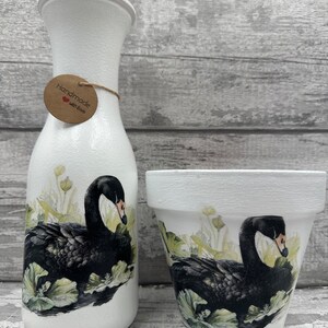 Könnte beinhalten: Ein Set aus zwei weißen Keramik-Pflanzgefäßen mit schwarzen Schwanen-Designs. Der größere Topf ist eine Vase mit einem braunen Etikett, auf dem "Handgefertigt mit Liebe" steht.
