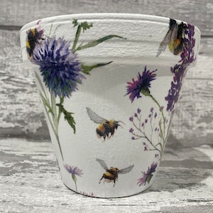 May include: White ceramic flower pot with a floral design featuring purple flowers and bees. The pot has a textured surface.