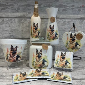 May include: A set of handmade home decor items featuring a German Shepherd dog design. The set includes a vase, a bottle, a jar, a heart-shaped plaque, and four coasters. All items are painted white with a watercolour-style design of a German Shepherd dog in a field of flowers. The items are labelled "Handmade with Love".