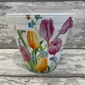 Peut inclure: Pot de fleurs en céramique blanche avec un motif floral représentant des tulipes roses et jaunes et des myosotis bleus.