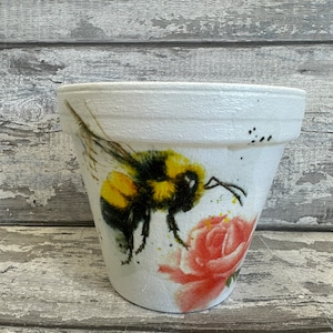 May include: White ceramic flower pot with a painted design of a bumblebee and a pink rose.