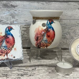 May include: A white ceramic wax melt warmer with a pheasant design. The warmer has a small heart charm hanging from it. A small white wax melt is shown in a clear container with a label that reads "Something Different" and "Made with love". A small tea light candle is also shown.