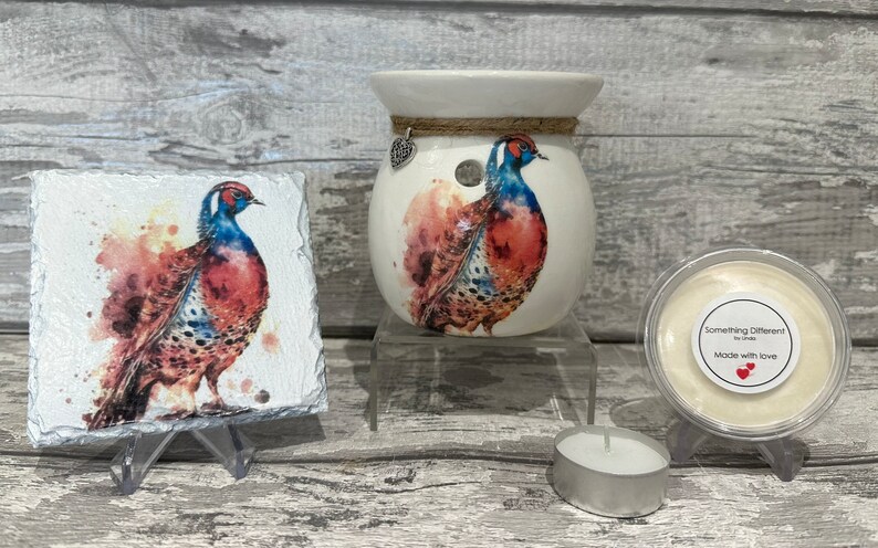 May include: A white ceramic wax melt warmer with a pheasant design. The warmer has a small heart charm hanging from it. A small white wax melt is shown in a clear container with a label that reads "Something Different" and "Made with love". A small tea light candle is also shown.