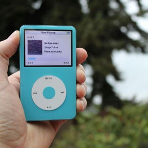 May include: A turquoise digital music player with a white control wheel and screen displaying "Now Playing." The screen shows song details: "Gethsemane," "Sleep Token," and "Even In Arcadia." The device is held in a hand against a blurred green background.