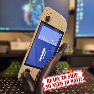 May include: A beige handheld gaming console with a black screen displaying the text Üdvözlünk! and a language selection menu. The device is supported by a dark gray, tentacle-shaped stand featuring visible suction cups on its curved surface.