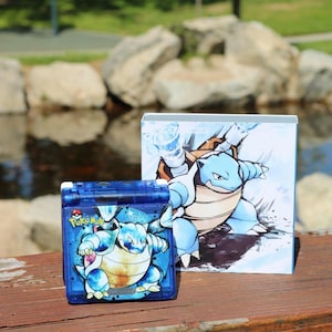 Op de afbeelding: Een blauwe handheld gameconsole met een Blastoise-afbeelding en het Pokemon-logo. Een bijpassende doos met een Blastoise-illustratie staat achter het apparaat. De items staan op een houten oppervlak met een vage achtergrond van water en rotsen.