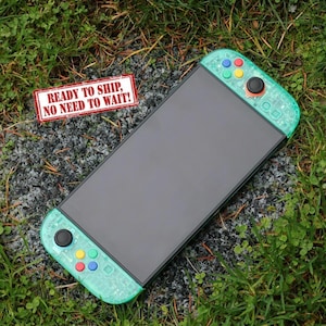 May include: A teal and black handheld gaming console with colorful buttons. The console has a large black screen and teal controllers on each side. The text "READY TO SHIP, NO NEED TO WAIT!" is on a small red and white sign.